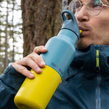A man standing outside in woodland and drinking from the Klean Kanteen 16oz Britanny kaleidoscope stainless steel reusable insulated rise bottle