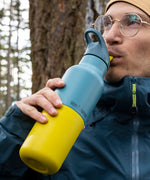 A man standing outside in woodland and drinking from the Klean Kanteen 16oz Britanny kaleidoscope stainless steel reusable insulated rise bottle