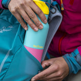 A adult hand putting the turquoise Kaleidoscope 16oz Klean Kanteen insulated rise bottle in a bottle holder on a backpack showing scale and the blue and pink colour way design with a yellow flip sports cap 