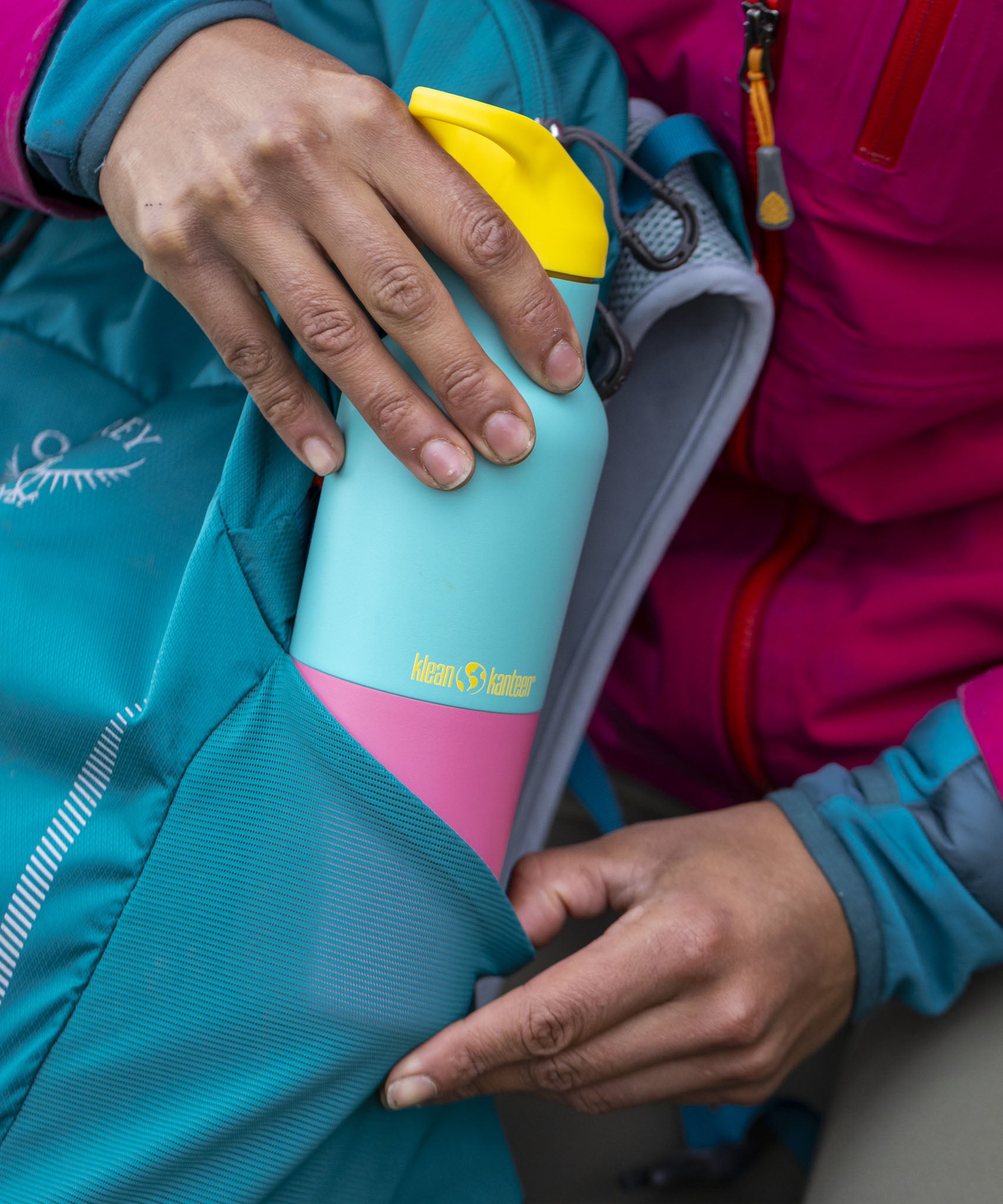 A adult hand putting the turquoise Kaleidoscope 16oz Klean Kanteen insulated rise bottle in a bottle holder on a backpack showing scale and the blue and pink colour way design with a yellow flip sports cap 