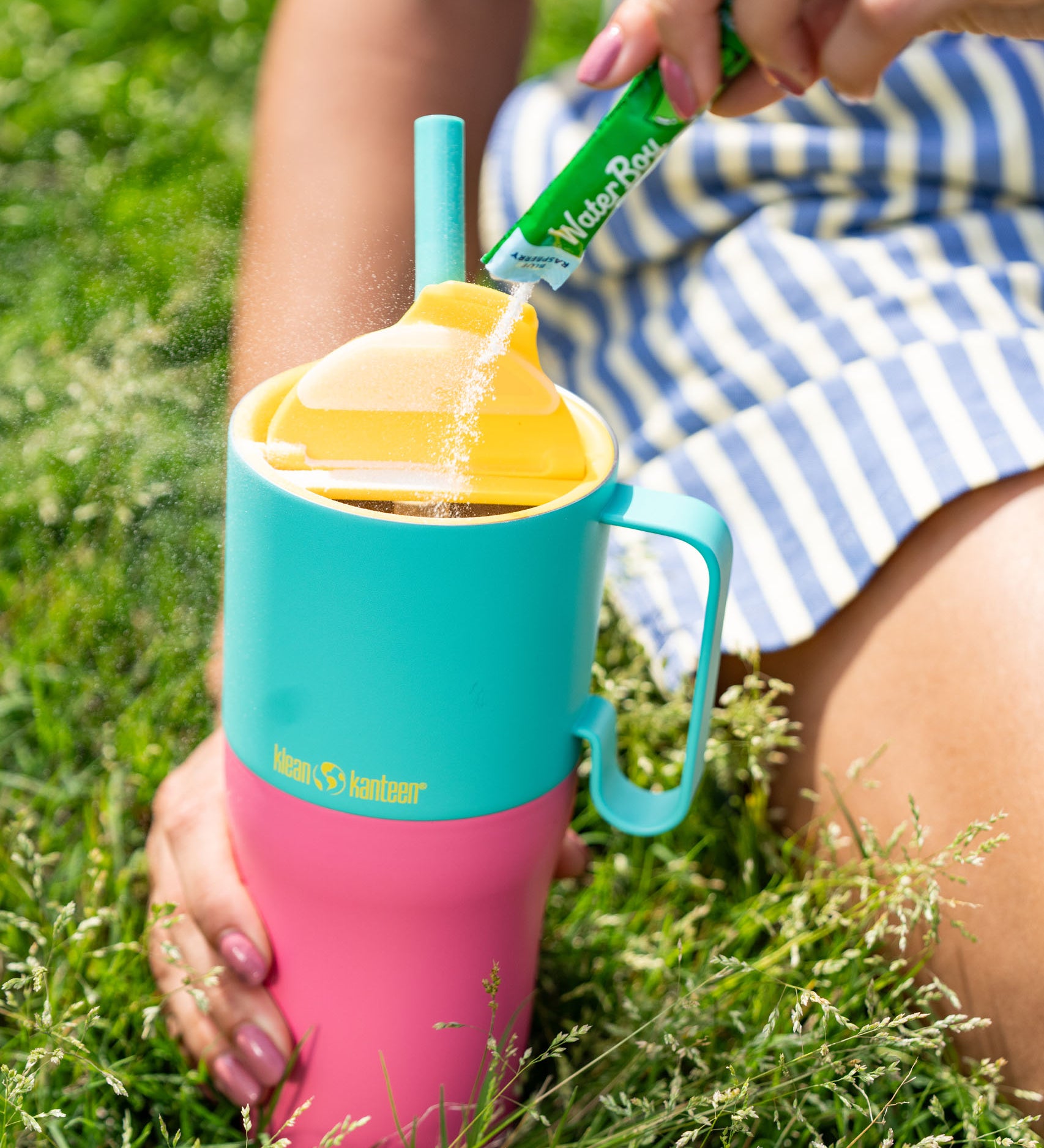 A close up of the Klean Kanteen 26oz turquoise stainless steel insulated rise tumbler on grass showing the yellow flip cap detail and the pink and turquoise colour way design