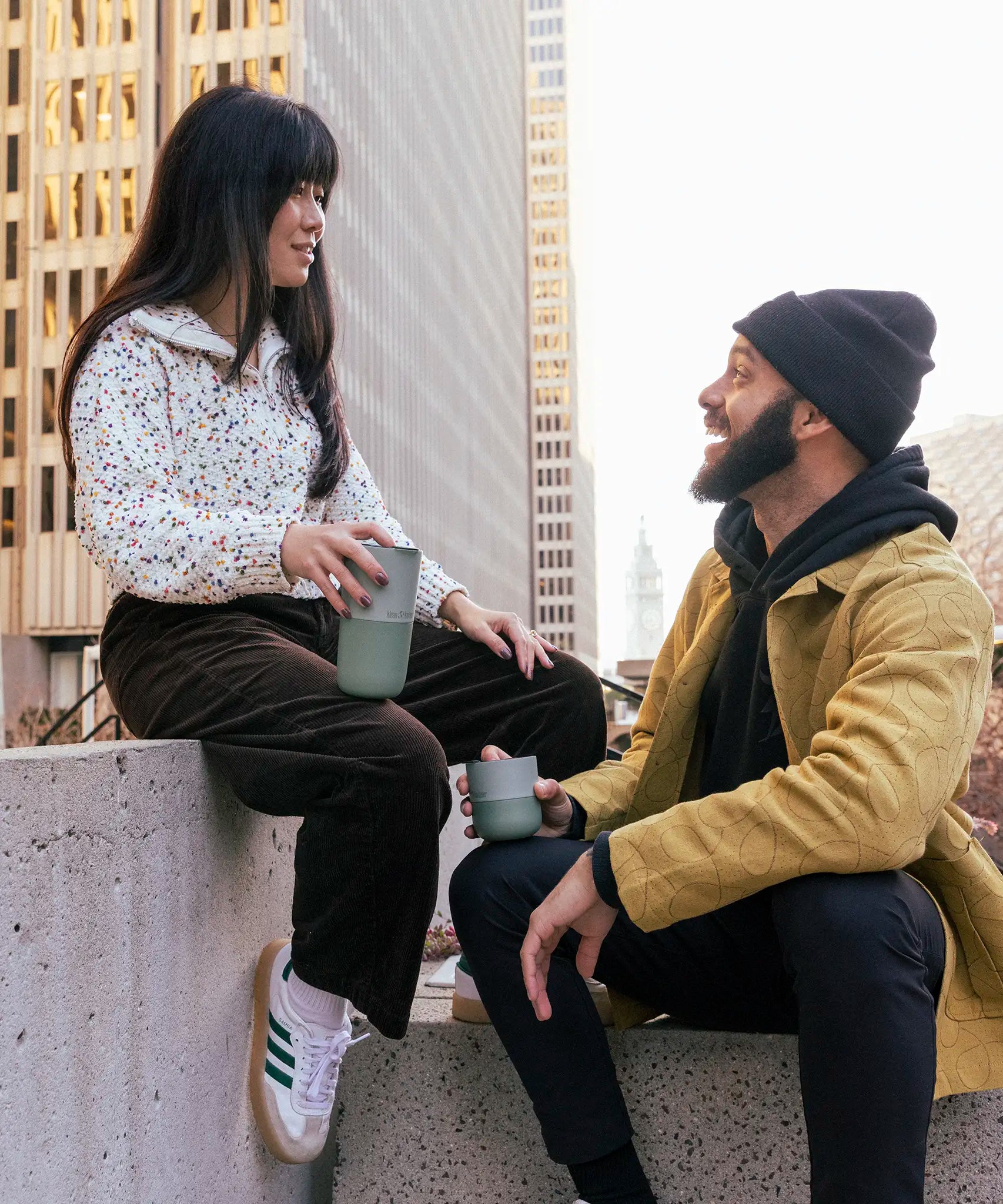 Adult holding a 16oz iceberg Klean Kanteen tumbler and another adult holding a 8oz lowball tumbler in the city