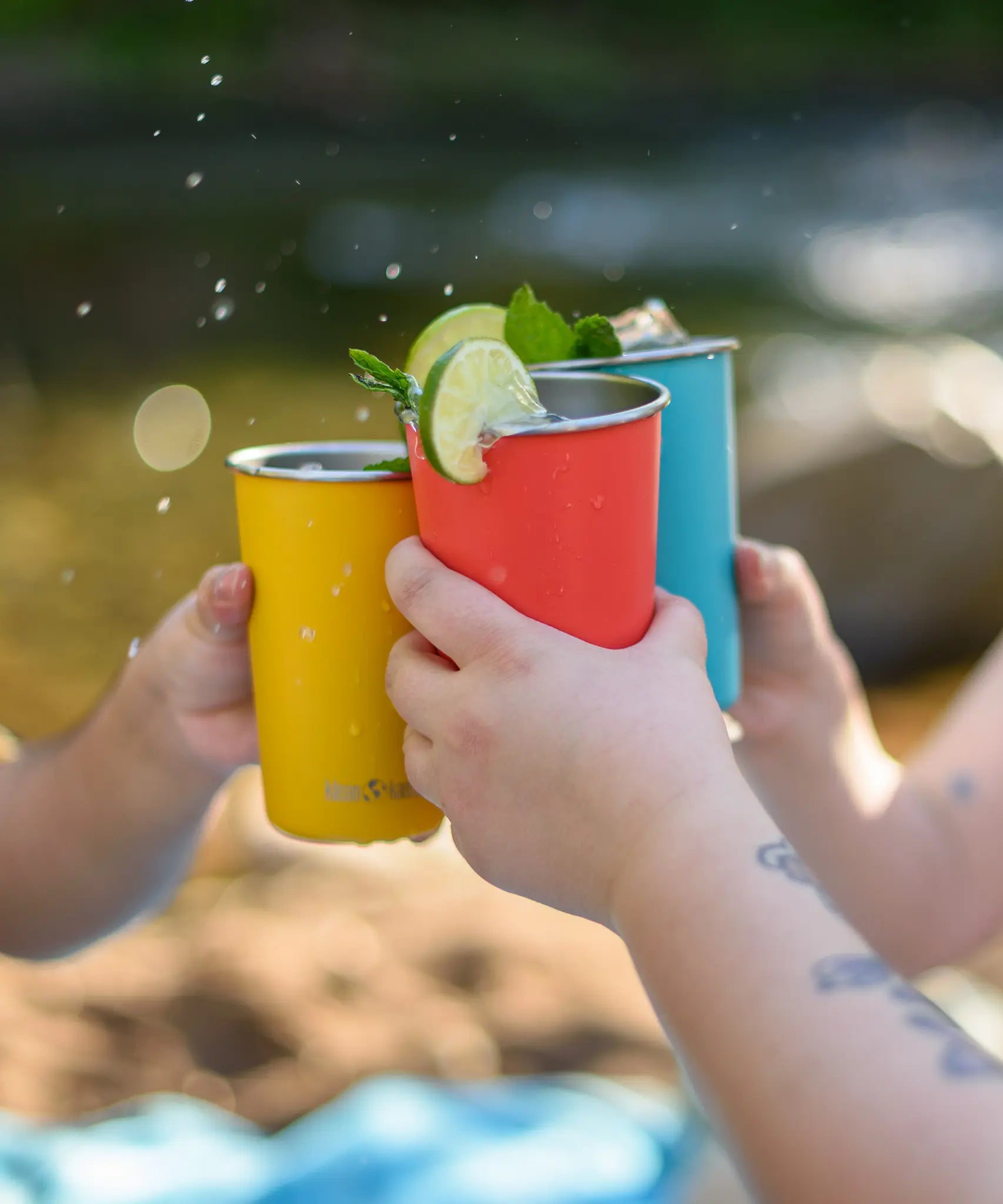 Klean Kanteen stainless steel 16oz pint cups in evening sky colours filled with drink and decorated with limes and garnish, three people making a cheering motion