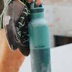 A climber holding the Klean Kanteen 20oz rise insulated Reusable Stainless Steel Water Bottle with loop cap in a Brittany blue colour. 