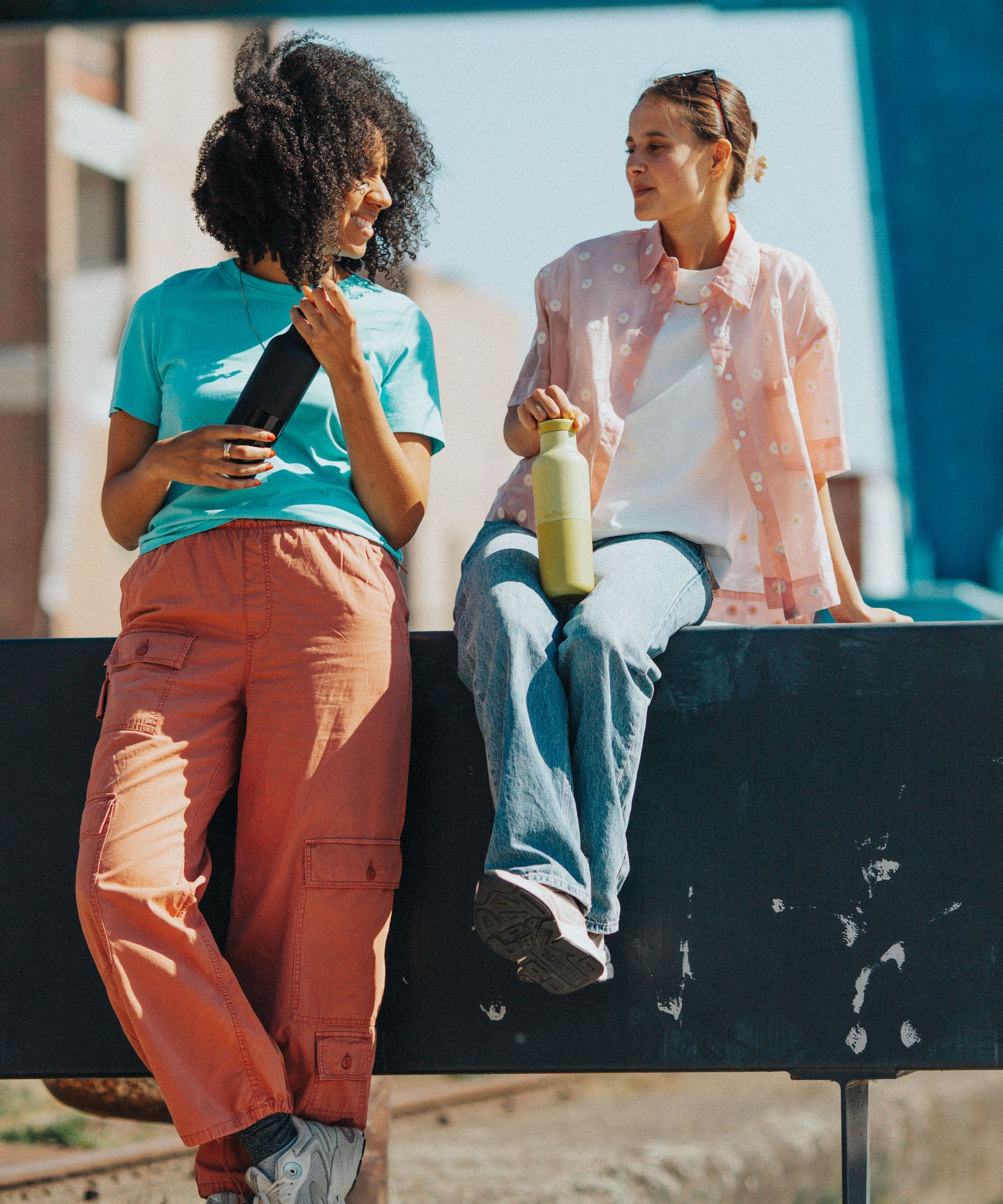 2 women sitting on a barrier and holding the 20oz klean Kanteen rise insulated classic bottles in black and acacia. A Reusable Stainless Steel Water Bottle with loop cap 