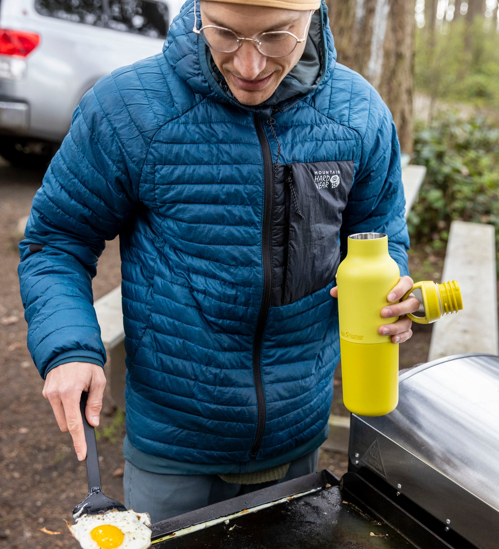 A man cooking outside and holding the Acacia 20oz rise Klean Kanteen water bottle with the loop cap off the bottle
