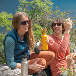 Two people sitting on rocks, one person is holding a Sunset yellow coloured Klean Kanteen 27oz Classic Narrow Flip Seal Sports Cap Reusable Stainless Steel Water Bottle there is another brushed steel bottle on the rock in front of them.