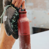 Adult holding a garnet red Klean Kanteen 20oz insulated bottle with matching lid and climbing shoes