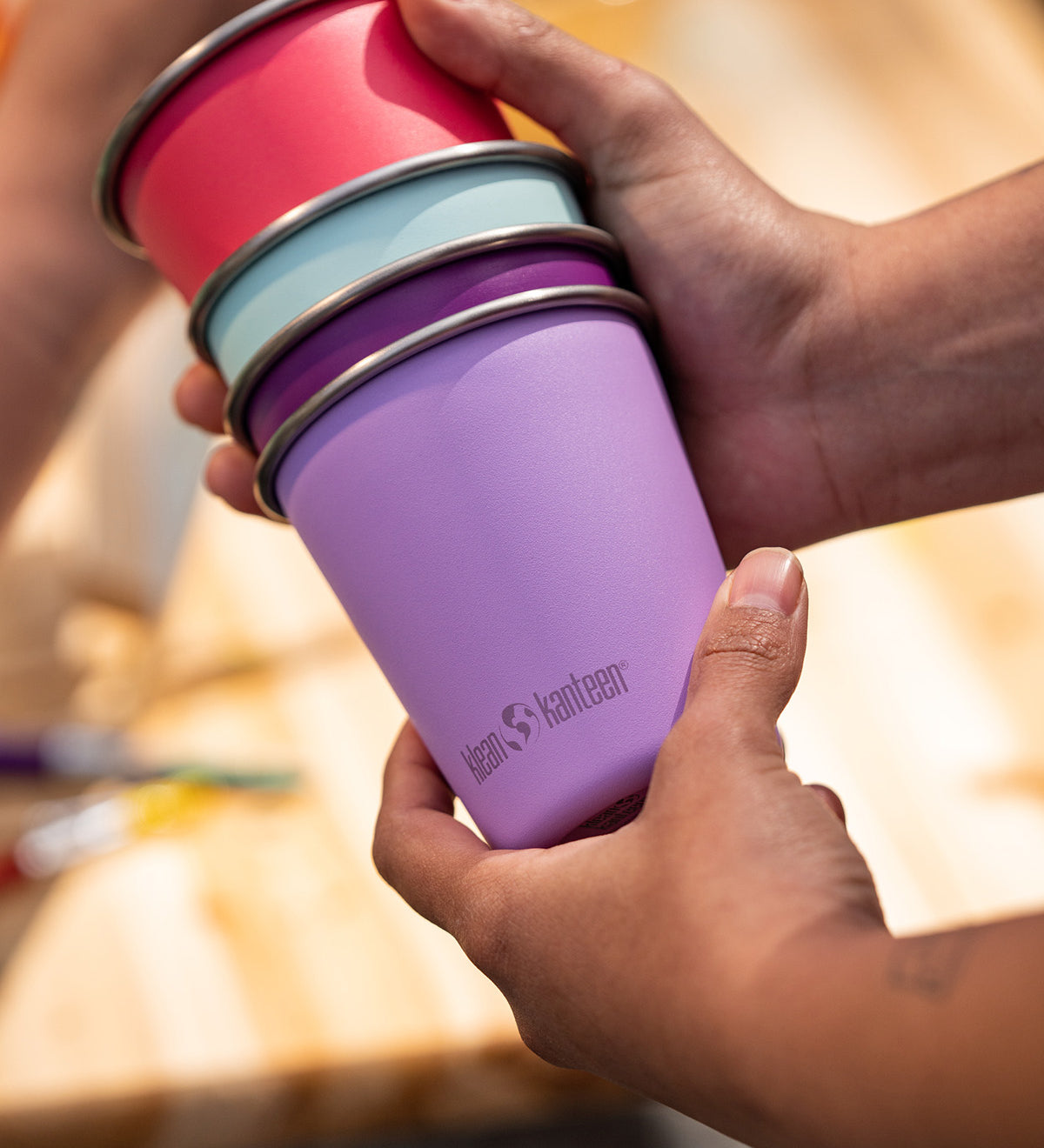Hands holding a stack of Klean Kanteen stainless steel kid cups 10oz in bright pinks and purples - plastic free, made from post-consumer steel, available to buy at Babipur.