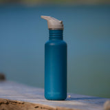 Blue Klean Kanteen bottle fitted with a taupe Flip Seal Sports Cap photographed beside water outdoors