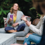 Close up of hand holding the Klean kanteen 10oz stainless steel cup in front of a woman sat on an outdoor seat