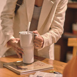 A close up of a person's hands holding a barley blue coloured 26oz handled rise tumbler from Klean Kanteen ready to lift it up off a table.