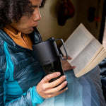 A woman drinking from a black Klean Kanteen 26oz handled Rise Tumbler whilst reading a book. 