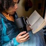 A woman drinking from a black Klean Kanteen 26oz handled Rise Tumbler whilst reading a book. 