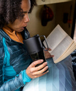 A woman drinking from a black Klean Kanteen 26oz handled Rise Tumbler whilst reading a book. 