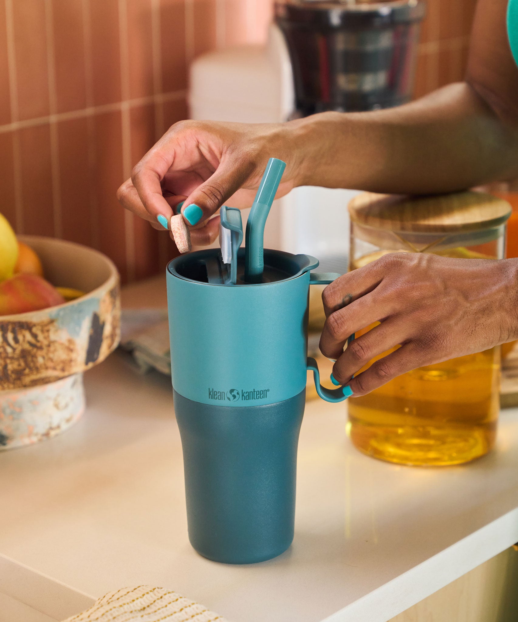 A close up of an adult's hands holding the Brittany Blue colourway Klean Kanteen handled, insulated Rise Tumbler in 26oz, preparing a drink on a kitchen work surface. 