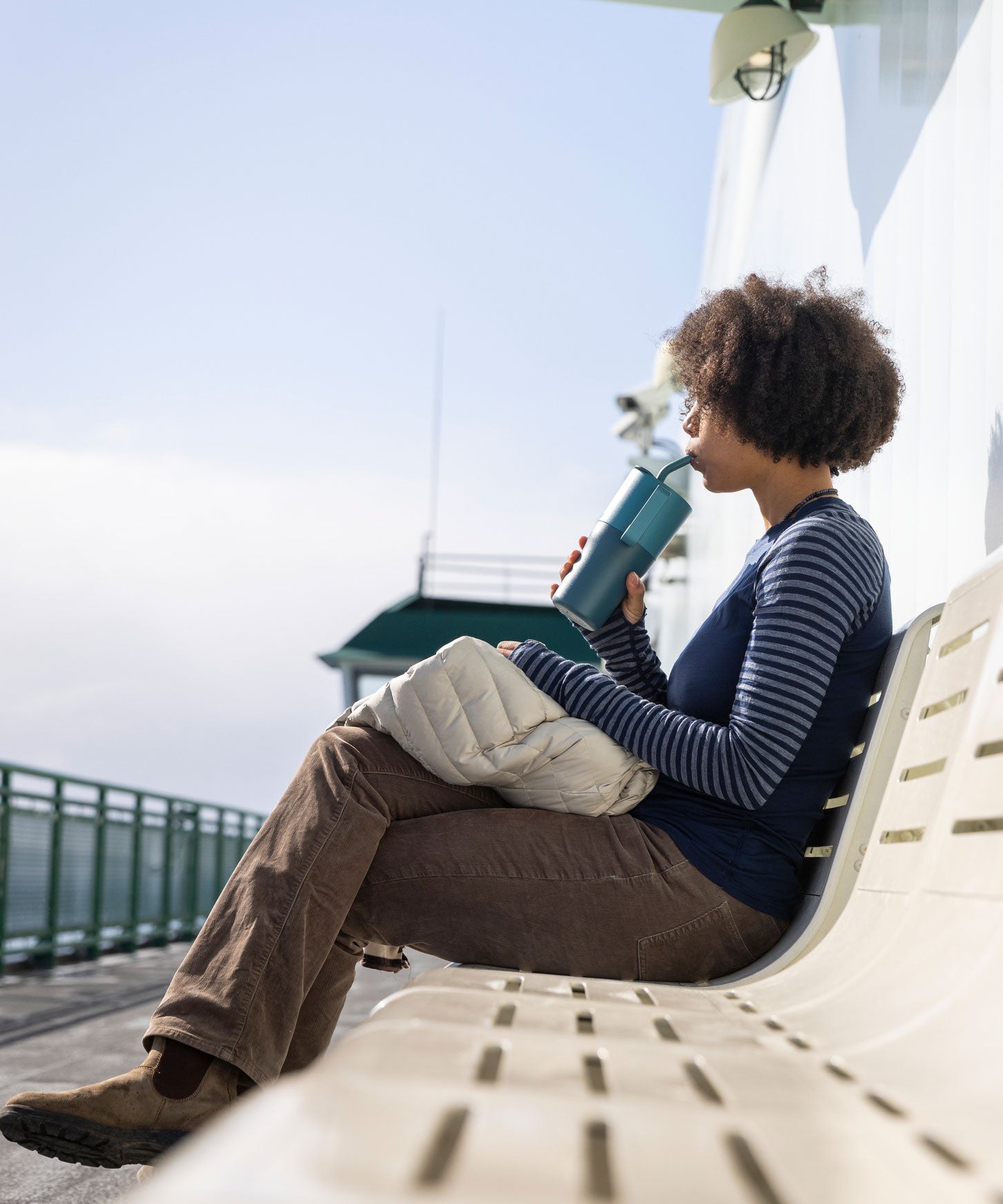 A woman sitting on a white bench sipping the straw on a brittany blue coloured 26oz Klean Kanteen rise handled tumbler. 