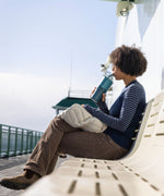A woman sitting on a white bench sipping the straw on a brittany blue coloured 26oz Klean Kanteen rise handled tumbler. 