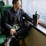 A man sitting by a window on a ferry. He is holding the base of a black coloured 26oz Klean Kanteen rise handled tumbler which is resting on the windowsill. 