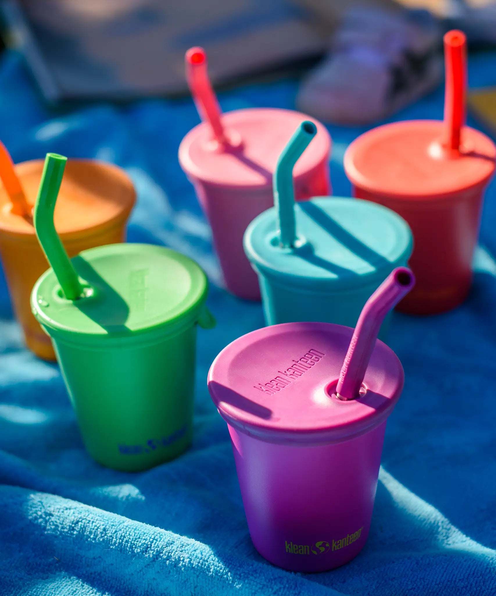 Six colours of Klean Kanteen 10oz/296ml Steel Kid Cups with Straw Lids placed on a blue blanket.