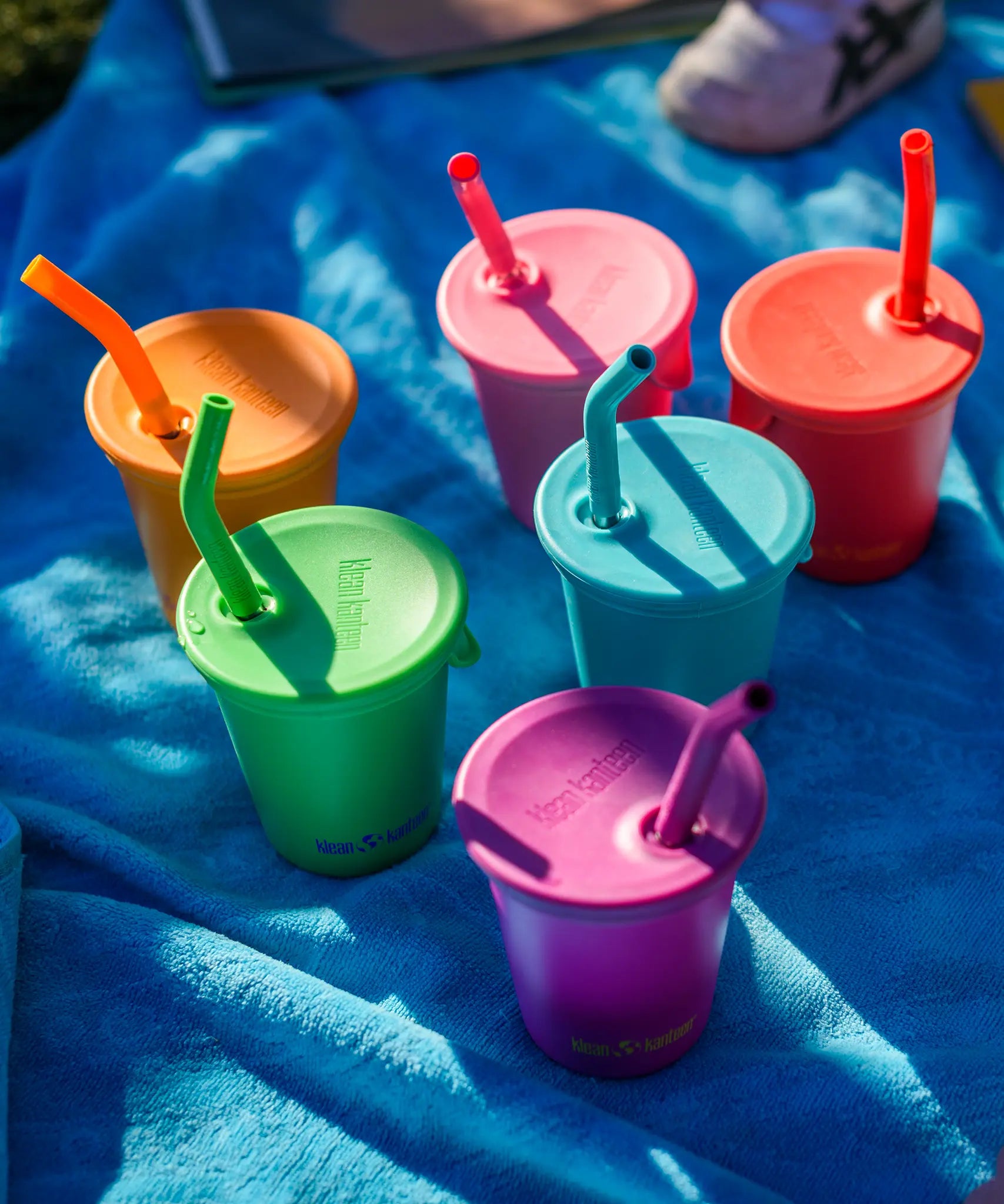 Six colours of Klean Kanteen 10oz/296ml Steel Kid Cups with Straw Lids placed on a blue blanket.