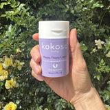 An adult hand holding the  tube of Kokoso organic coconut oil Happy Nappy Cream with a flowery hedge in the background available at Babipur. 