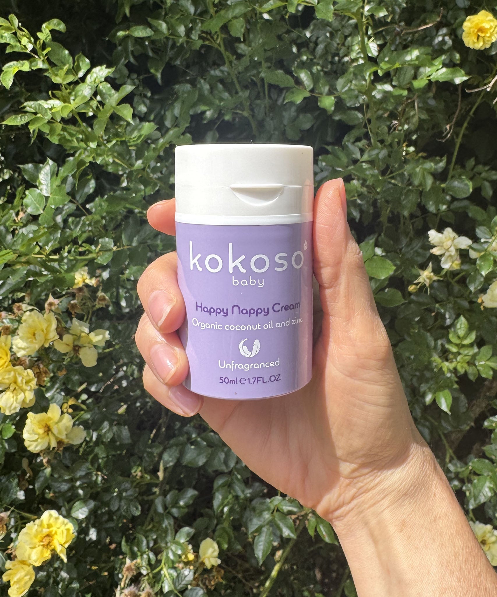 An adult hand holding the  tube of Kokoso organic coconut oil Happy Nappy Cream with a flowery hedge in the background available at Babipur. 