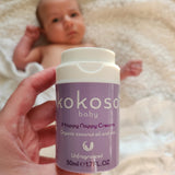 An adult hand holding the  tube of Kokoso organic coconut oil Happy Nappy Cream above a baby. Kokoso's range of organic  coconut oil baby care is available at Babipur. 