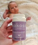 An adult hand holding the  tube of Kokoso organic coconut oil Happy Nappy Cream above a baby. Kokoso's range of organic  coconut oil baby care is available at Babipur. 