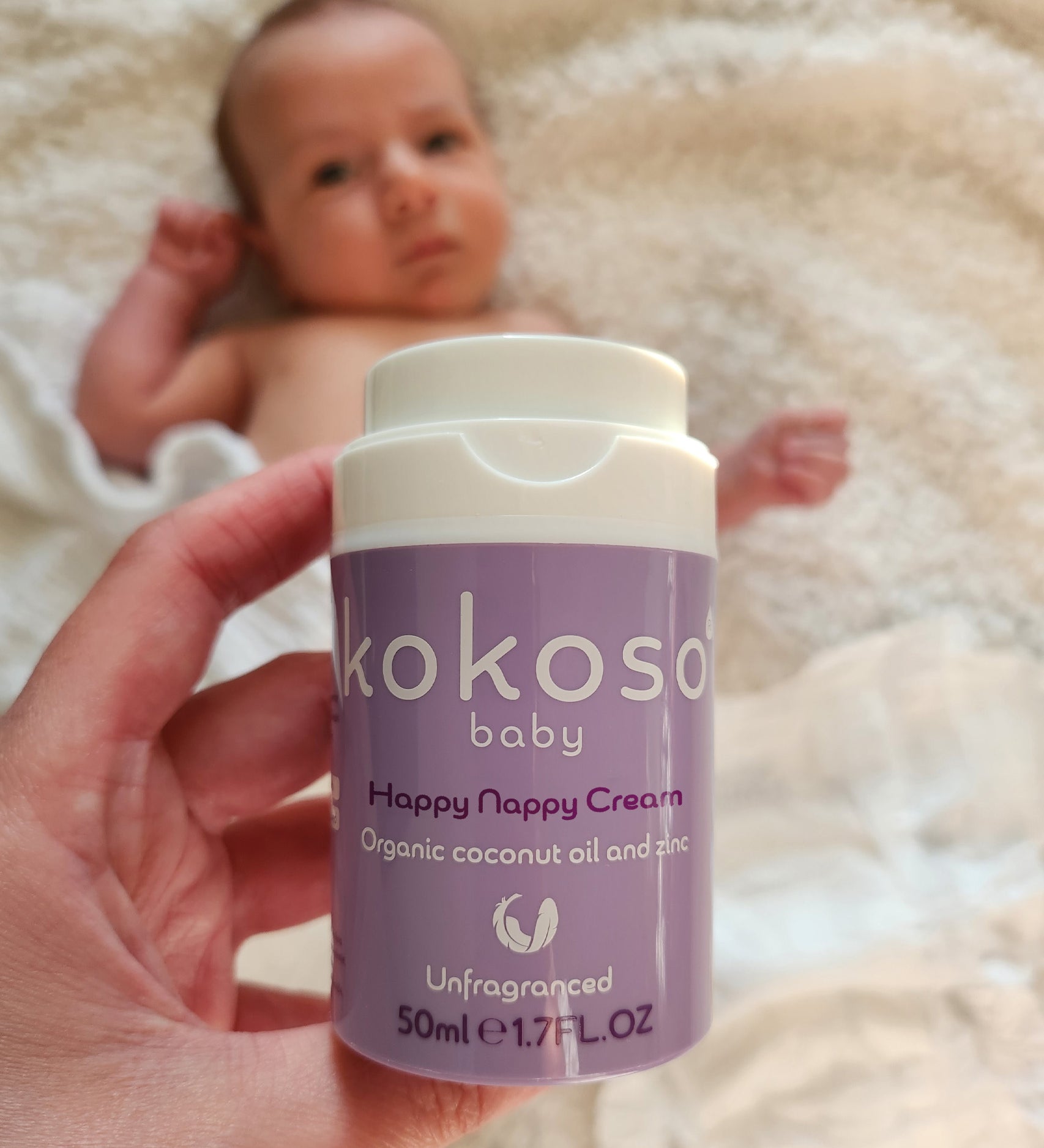 An adult hand holding the  tube of Kokoso organic coconut oil Happy Nappy Cream above a baby. Kokoso's range of organic  coconut oil baby care is available at Babipur. 