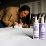 Close up of the Kokoso coconut oil lotion bottles on a white bed in front of a woman and a baby