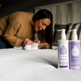 Close up of the Kokoso coconut oil lotion bottles on a white bed in front of a woman and a baby