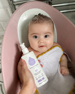 A child sat in a baby bath with a person holding the Kokoso Fragrance Free Hair and Body Wash bottle next to them
