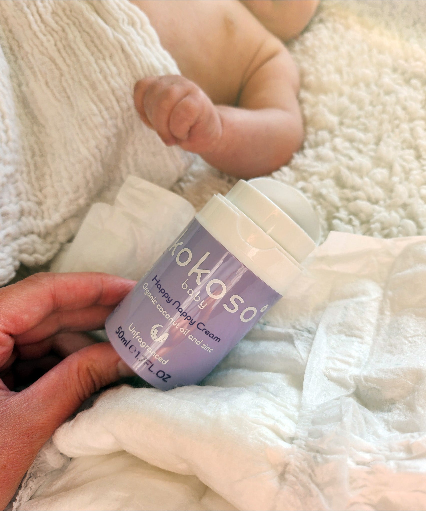 An adult hand holding the tube of Kokoso organic coconut oil Happy Nappy Cream next to a lying baby.  Kokoso's range of organic coconut oil baby care is available at Babipur. 