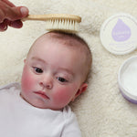Close up of baby laying on a white fluffy background having its hair brushed with the Kokoso natural wooden hairbrush