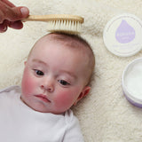 Close up of baby laying on a white fluffy background having its hair brushed with the Kokoso natural wooden hairbrush