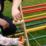 The Kraul DIY Basket Cable Car STEM Kit set up outdoors attached to a TriClimb.  An adult's hand can be seen turning the wheel control in the background. 