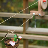 A wicker basket with a green passenger felt doll and pink petals travelling on the Kraul DIY Basket Cable Car STEM Kit. 