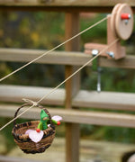 A wicker basket with a green passenger felt doll and pink petals travelling on the Kraul DIY Basket Cable Car STEM Kit. 