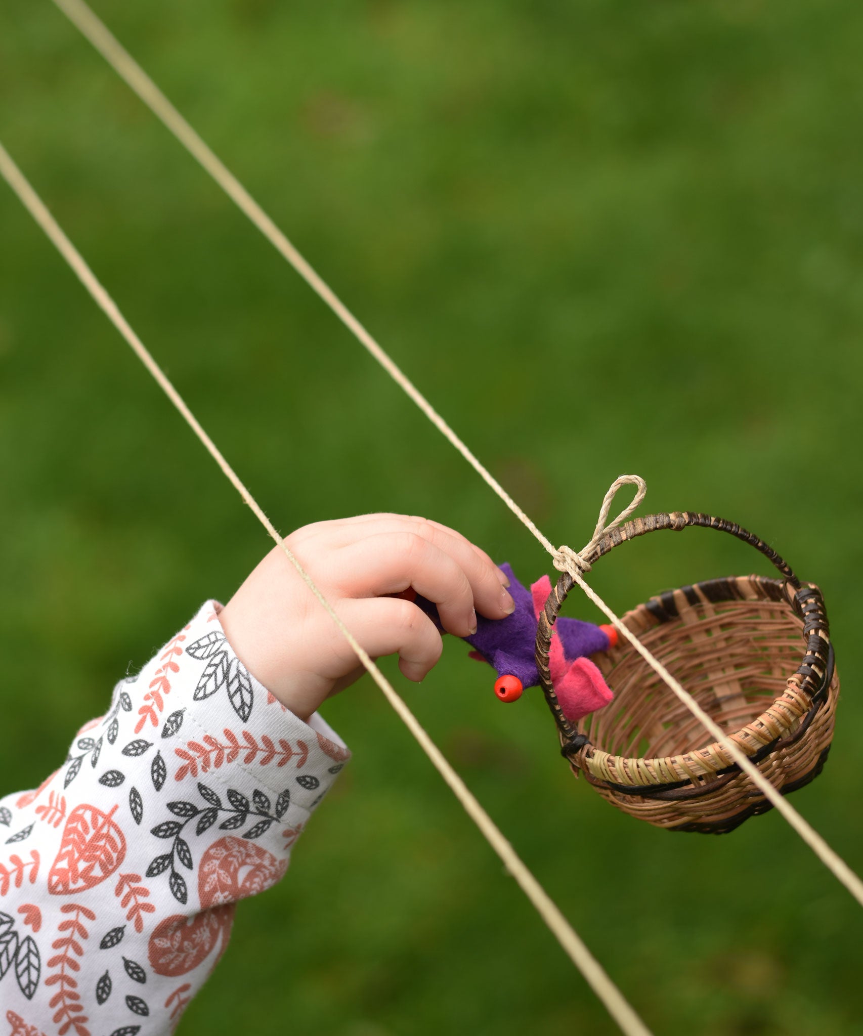A child's hand placing a felt passenger doll into a wicker basket from the Kraul DIY Basket Cable Car STEM Kit.  An adult's hand can be seen turning the wheel control in the background. 
