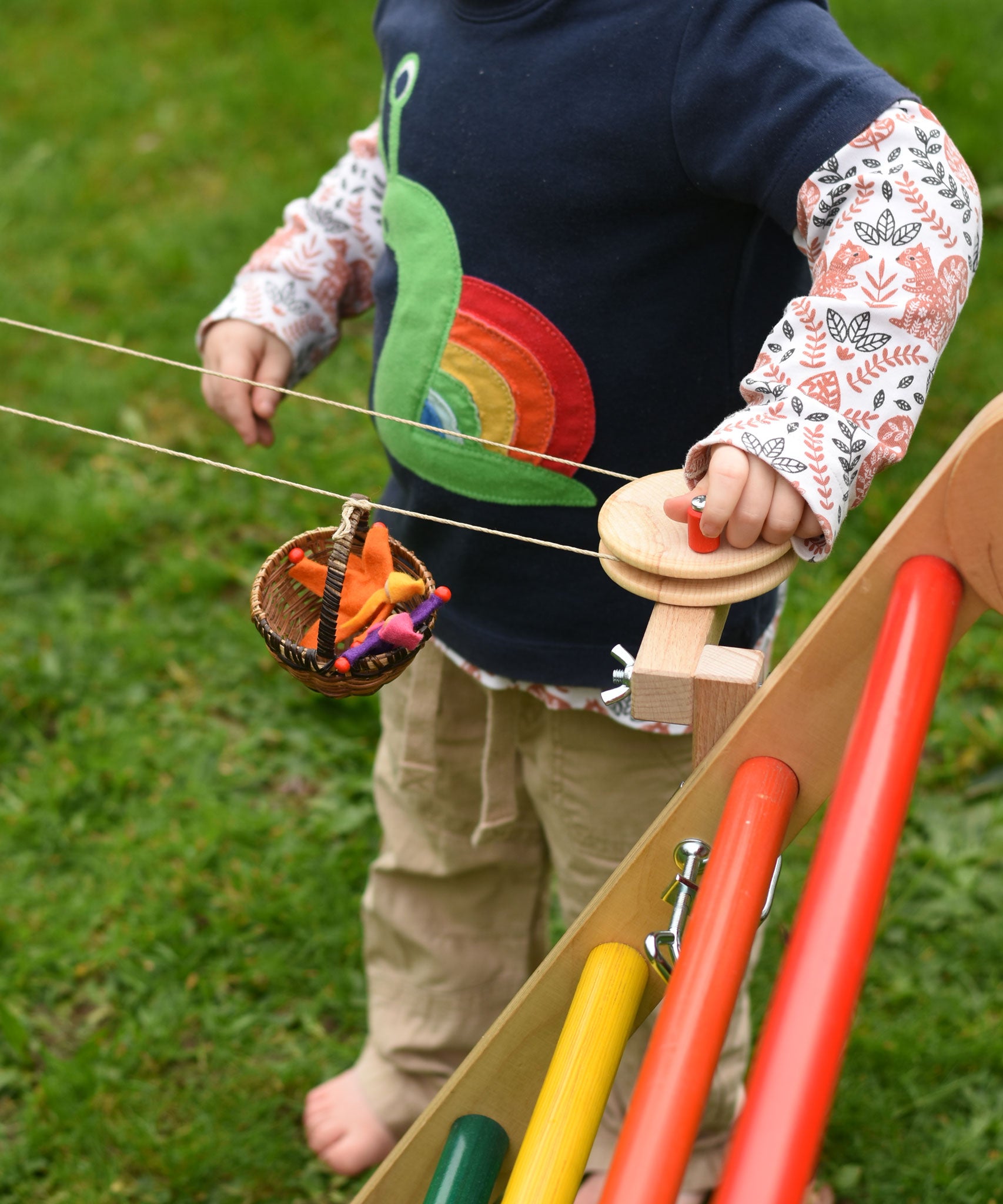 A close up of a child's hand turning the wheel of the Kraul DIY Basket Cable Car STEM Kit as a basket filled with felt passenger moves along the string.