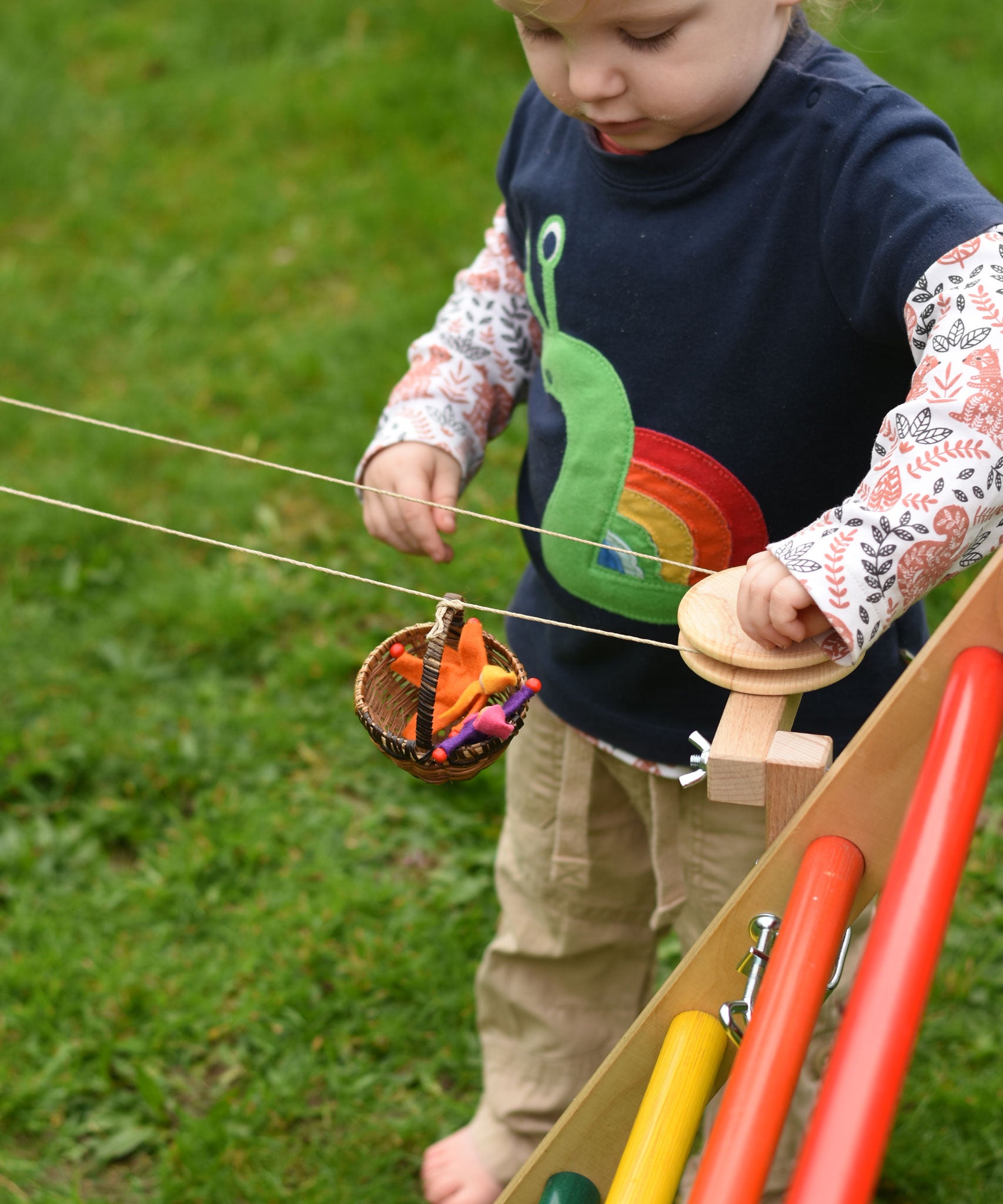 A close up of a child's hand turning the wheel of the Kraul DIY Basket Cable Car STEM Kit as a basket filled with felt passenger moves along the string.