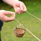 A close up of an adult's hand holding the string and attaching a wicker basket to the Kraul DIY Basket Cable Car STEM Kit. 