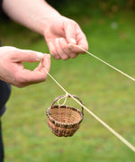 A close up of an adult's hand holding the string and attaching a wicker basket to the Kraul DIY Basket Cable Car STEM Kit. 
