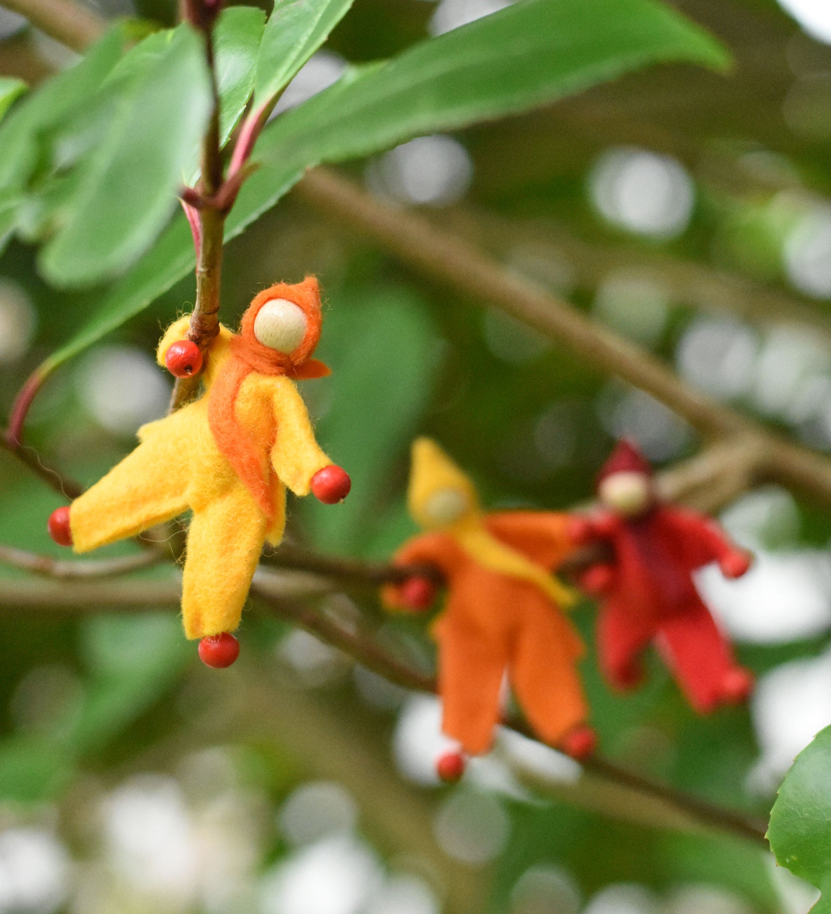 A yellow felt Kraul Little Passenger Doll handing on a little branch on a tree. Other dolls can be seen in the background. These pose-able dolls are perfect to fit inside smaller toy vehicles and for use with the Kraul cable cars.