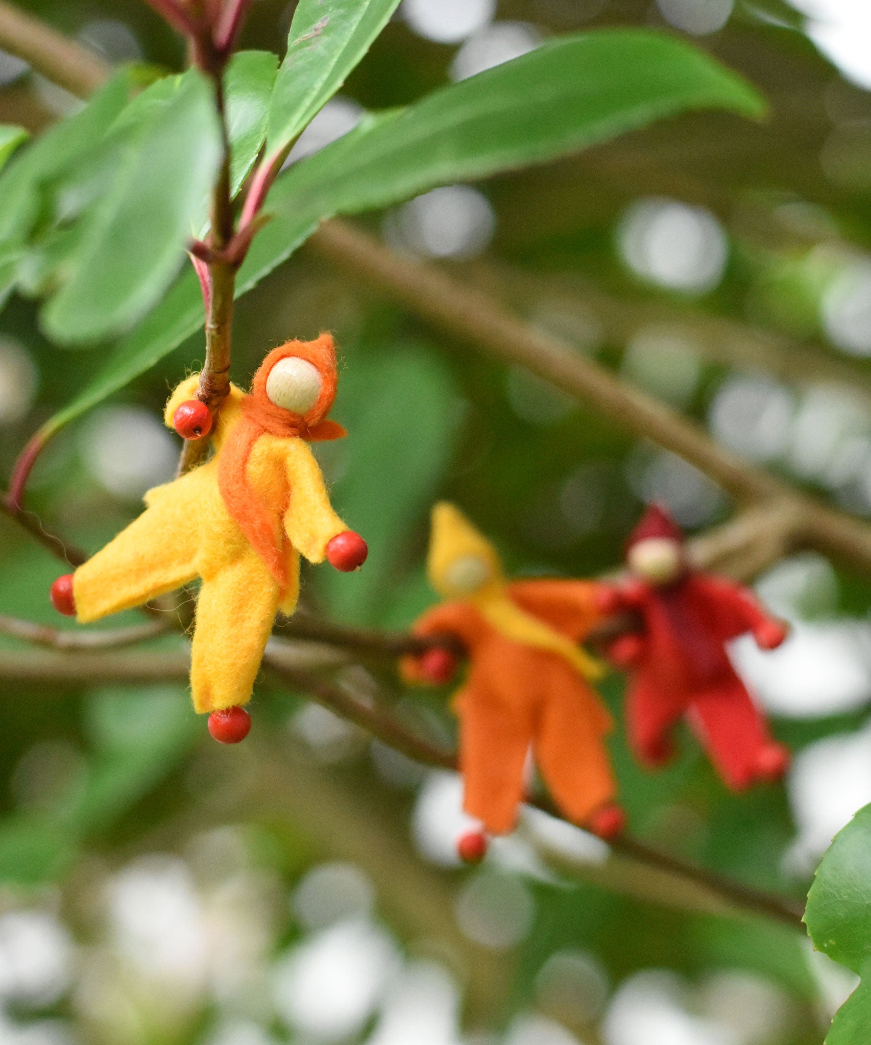 A yellow felt Kraul Little Passenger Doll handing on a little branch on a tree. Other dolls can be seen in the background. These pose-able dolls are perfect to fit inside smaller toy vehicles and for use with the Kraul cable cars.
