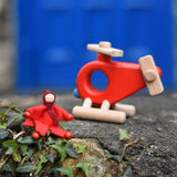 A red felt Kraul Little Passenger Doll sitting on a wall in front of a Bajo fire engine helicopter. These pose-able dolls are perfect to fit inside smaller toy vehicles and for use with the Kraul cable cars.