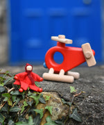 A red felt Kraul Little Passenger Doll sitting on a wall in front of a Bajo fire engine helicopter. These pose-able dolls are perfect to fit inside smaller toy vehicles and for use with the Kraul cable cars.