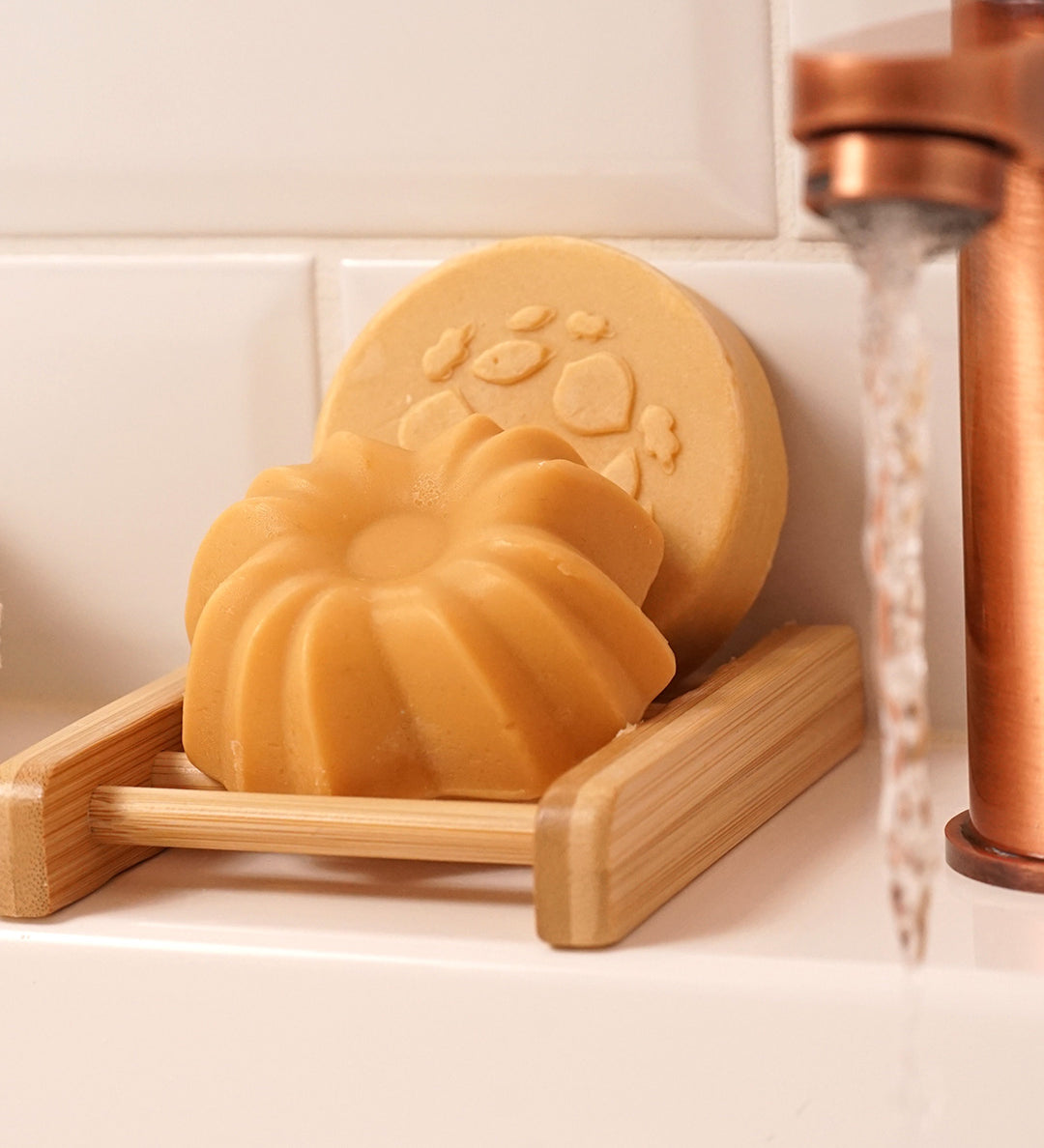 Lamazuna Keratin Shine solid conditioner bar resting in a wooden soap dish with the Keratin Repair shampoo bar behind it beside a running tap