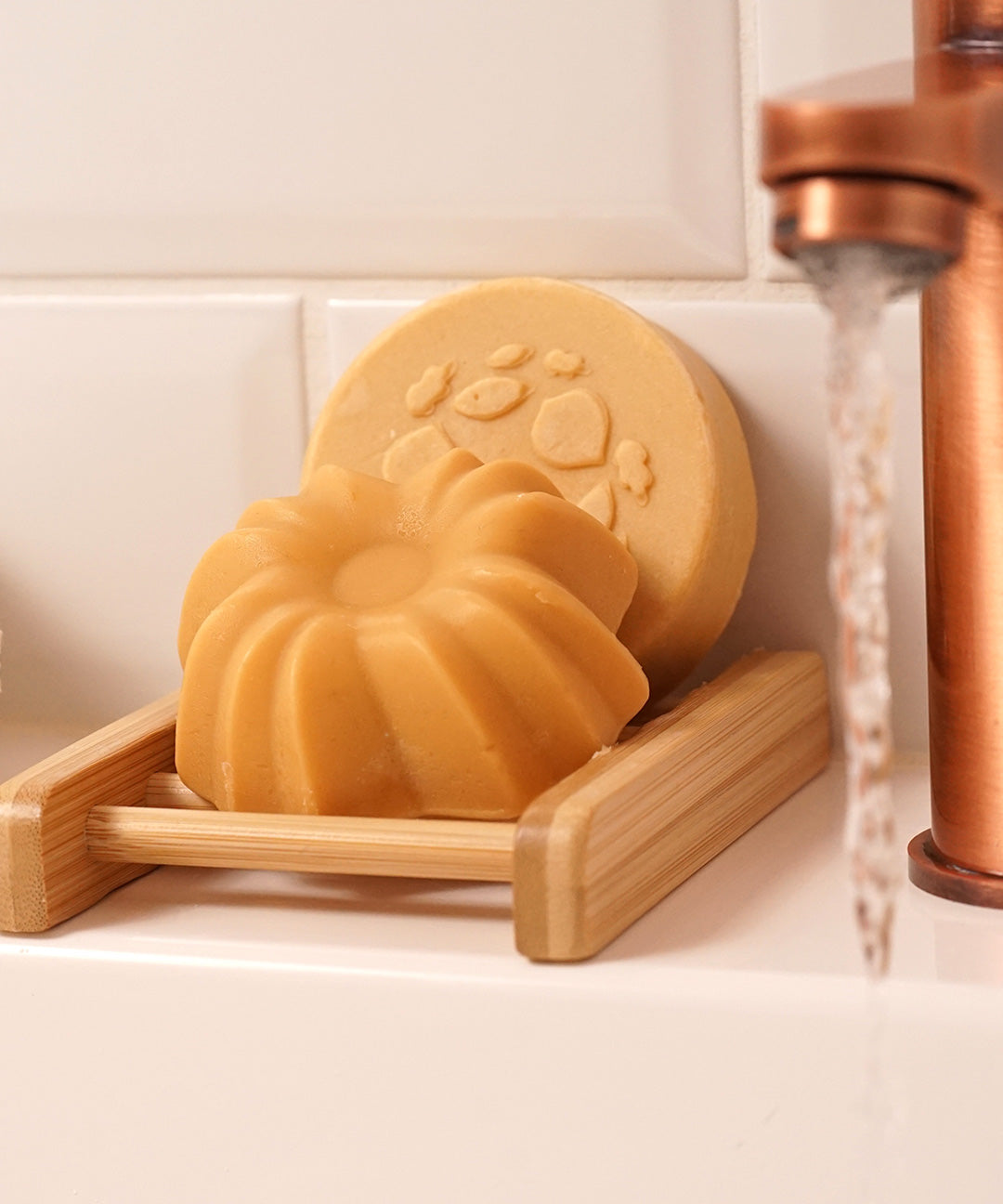 Lamazuna Keratin Shine solid conditioner bar resting in a wooden soap dish with the Keratin Repair shampoo bar behind it beside a running tap